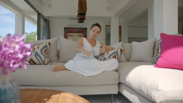 Young lady in white dress sits in hotel living room on large sofa with pillows drinks coffee and reads magazine, pleasant holiday concept