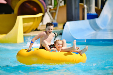 Boy and girl have fun on water slide in outdoor aquapark. Little children floating on yellow...