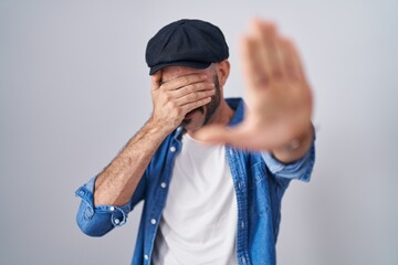 Hispanic man with beard standing over isolated background covering eyes with hands and doing stop...