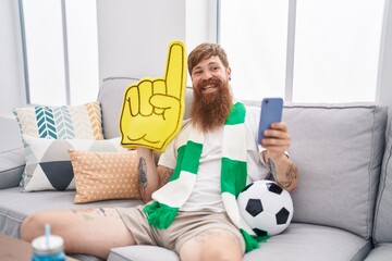 Young redhead man supporting soccer team using smartphone holding big hand at home