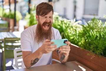 Young redhead man watching video on smartphone sitting on table at coffee shop terrace