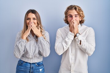 Young couple standing over blue background laughing and embarrassed giggle covering mouth with...