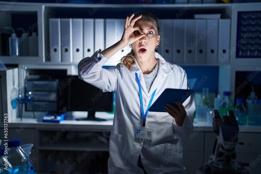 Poster Beautiful blonde woman working at scientist laboratory late at night doing ok gesture shocked with surprised face, eye looking through fingers. unbelieving expression.