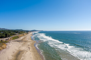 Aerial view of Beverly Beach on the Oregon Coast with gentle breaking waves on the sandy beach, the Yaquina Head Lighthouse and beach goers in the distance and a bright blue sky with copy space.