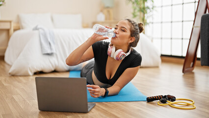 Young beautiful hispanic woman wearing sportswear lying on yoga mat drinking water at bedroom