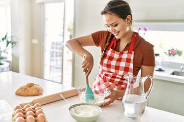 Young beautiful hispanic woman smiling confident mixing water and flour on bowl at the kitchen