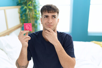 Young hispanic man sitting on the bed holding condom serious face thinking about question with hand...