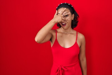 Young hispanic woman standing over red background peeking in shock covering face and eyes with...