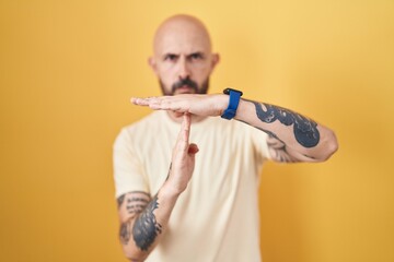 Hispanic man with tattoos standing over yellow background doing time out gesture with hands, frustrated and serious face