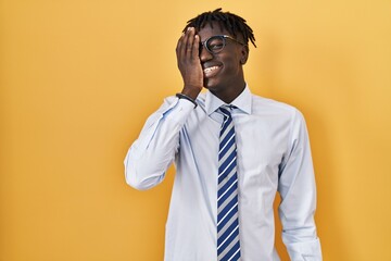African man with dreadlocks standing over yellow background covering one eye with hand, confident smile on face and surprise emotion.