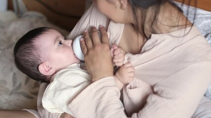 Sitting on the bed, a young mother feeds her baby with a specialized formula, demonstrating the diverse choices parents make today, sometimes opting for or relying on alternative feeding methods.