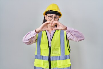 Hispanic girl wearing builder uniform and hardhat smiling in love doing heart symbol shape with hands. romantic concept.