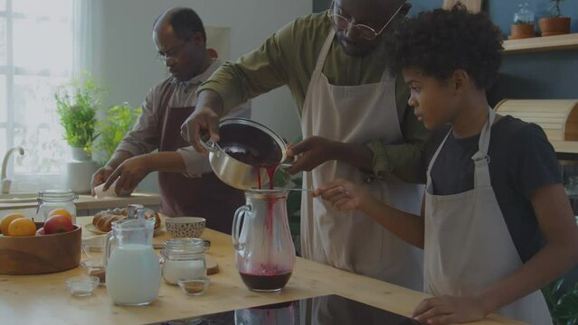 African American father and little son straining Zobo tea into glass pitcher as their grandfather serving Meskouta cake while preparing traditional food for home family dinner