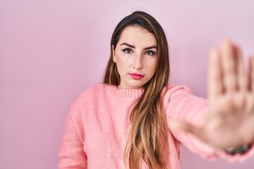 Young hispanic woman standing over pink background doing stop sing with palm of the hand. warning expression with negative and serious gesture on the face.