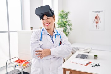 Young hispanic doctor woman wearing doctor uniform and virtual reality glasses winking looking at...