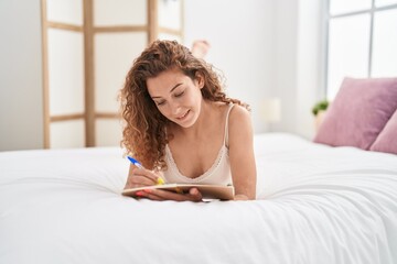 Young beautiful hispanic woman writing on notebook lying on bed at bedroom