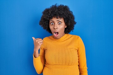 Fototapeta na wymiar Black woman with curly hair standing over blue background surprised pointing with hand finger to the side, open mouth amazed expression.