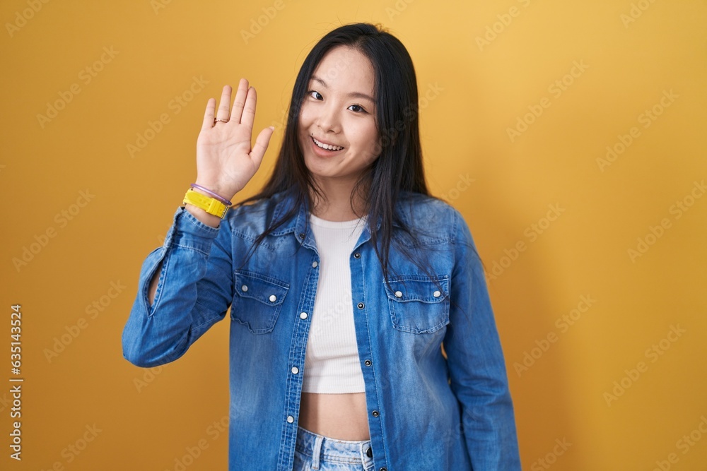 Sticker young asian woman standing over yellow background waiving saying hello happy and smiling, friendly w