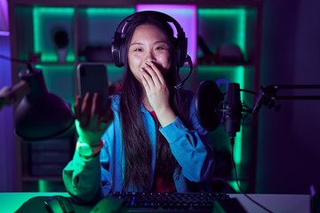 Young asian woman playing video games with smartphone laughing and embarrassed giggle covering mouth with hands, gossip and scandal concept