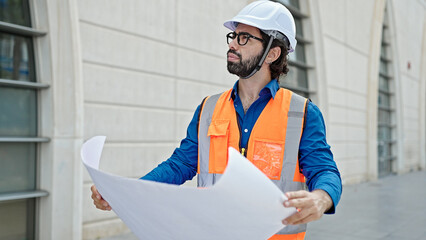 Young hispanic man architect holding blueprints looking around at construction place