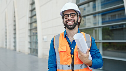 Young hispanic man architect smiling confident holding blueprints at construction place