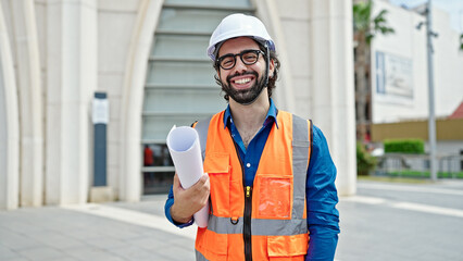 Young hispanic man architect smiling confident holding blueprints at construction place