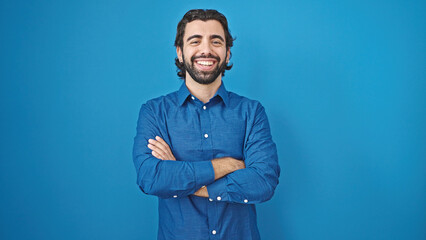 Young hispanic man smiling confident standing with arms crossed gesture over isolated blue...