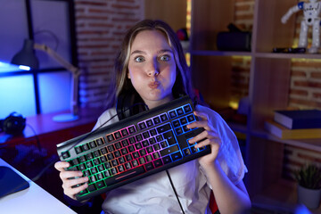 Young caucasian woman holding gamer keyboard puffing cheeks with funny face. mouth inflated with...