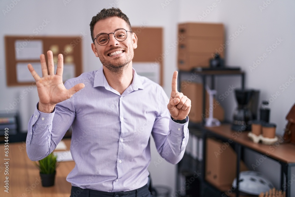 Wall mural Young hispanic man at the office showing and pointing up with fingers number six while smiling confident and happy.