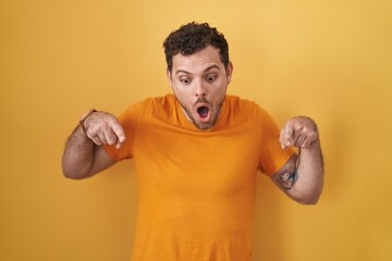 Young hispanic man standing over yellow background pointing down with fingers showing advertisement, surprised face and open mouth