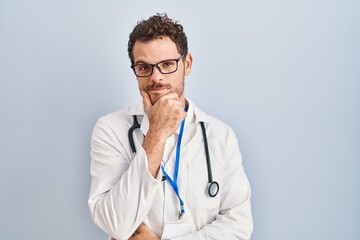 Young hispanic man wearing doctor uniform and stethoscope looking confident at the camera with smile with crossed arms and hand raised on chin. thinking positive.