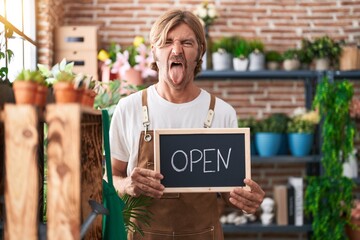 Caucasian man with mustache working at florist holding open sign sticking tongue out happy with funny expression.