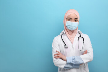 Muslim woman wearing hijab, medical uniform and protective mask on light blue background, space for text