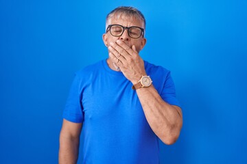 Hispanic man with grey hair standing over blue background bored yawning tired covering mouth with hand. restless and sleepiness.