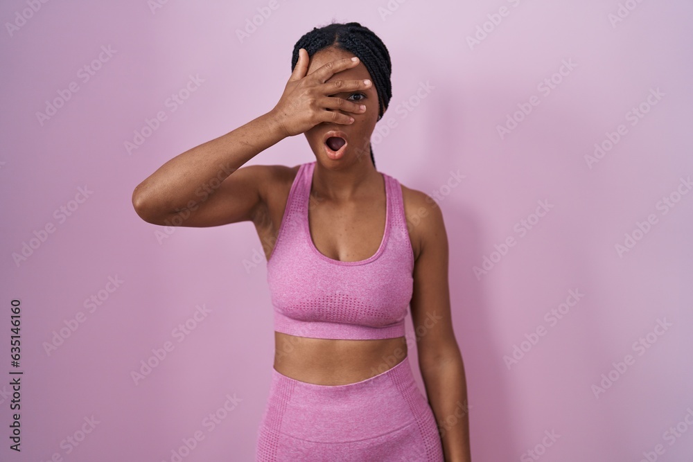 Poster African american woman with braids wearing sportswear over pink background peeking in shock covering face and eyes with hand, looking through fingers with embarrassed expression.