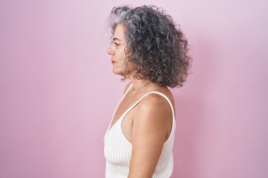 Middle age woman with grey hair standing over pink background looking to side, relax profile pose with natural face and confident smile.