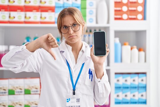 Young caucasian woman working at pharmacy drugstore showing smartphone screen with angry face, negative sign showing dislike with thumbs down, rejection concept