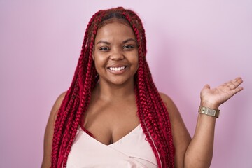 African american woman with braided hair standing over pink background smiling cheerful presenting and pointing with palm of hand looking at the camera.