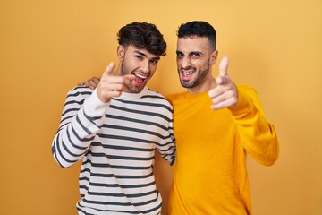 Young hispanic gay couple standing over yellow background pointing fingers to camera with happy and funny face. good energy and vibes.