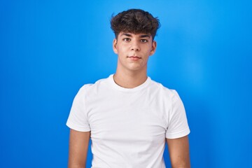 Hispanic teenager standing over blue background relaxed with serious expression on face. simple and...