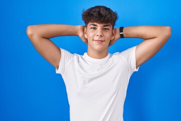Hispanic teenager standing over blue background relaxing and stretching, arms and hands behind head and neck smiling happy