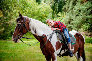 little handsome blonde smiling boy in red checkered shirt  riding horse in green forest on sunny day
