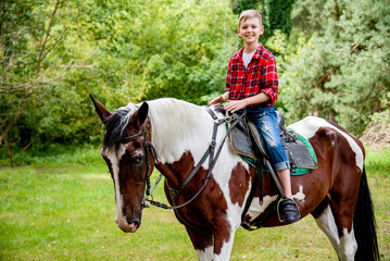 little handsome blonde smiling boy in red checkered shirt  riding horse in green forest on sunny day