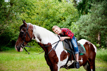 little handsome blonde smiling boy in red checkered shirt  riding horse in green forest on sunny day