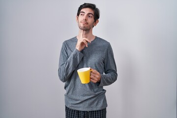 Young hispanic man wearing pajama drinking a cup of coffee thinking concentrated about doubt with...