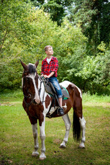 Fototapeta premium little handsome blonde smiling boy in red checkered shirt riding horse in green forest on sunny day