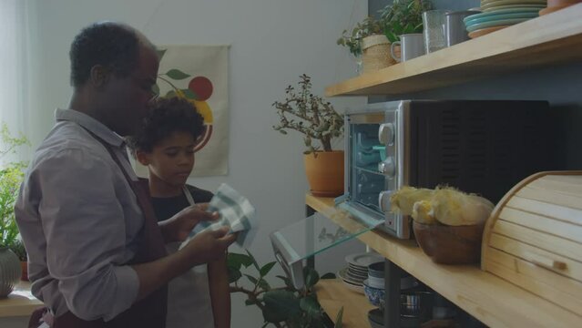 African American grandfather in apron taking off baked cake in silicone mold while cooking together with little grandson in kitchen at home