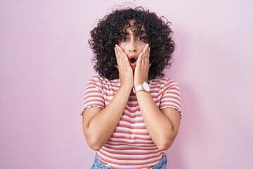 Young middle east woman standing over pink background afraid and shocked, surprise and amazed expression with hands on face