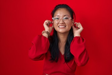 Asian young woman standing over red background smiling pulling ears with fingers, funny gesture....