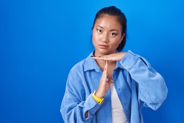 Asian young woman standing over blue background doing time out gesture with hands, frustrated and serious face